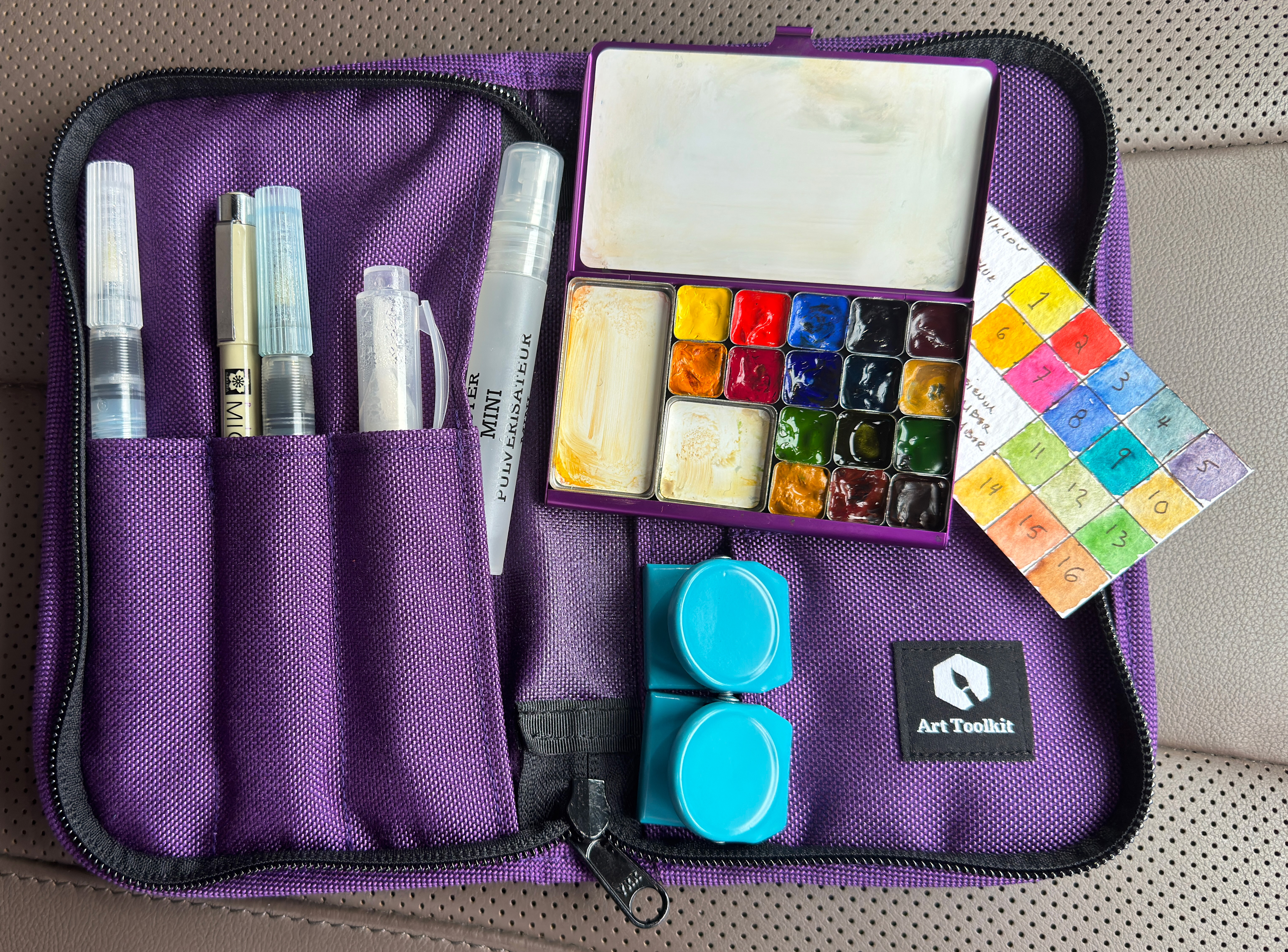 A small and thin watercolor palette with 16 small pans for colors, and 2 mixing wells, a swatch card of all the colors, on top of a small purple zip up pouch. The pouch is branded Art Toolkit. There are waterbrushes and pens in pen slots. And two magnetic fridge clips