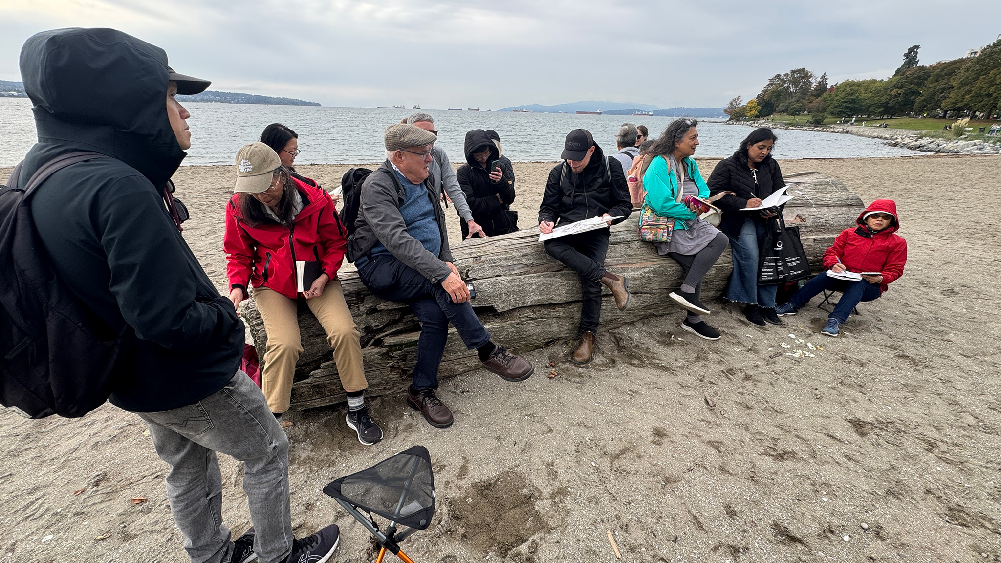 Felix Scheinberger sitting on a large deadwood log, with his sketchbook on his lap. There are many people around him, some sitting on the log, others standing behind or in front of him.